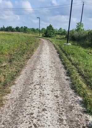 Narrow dirt road with tire marks winding through grassy fields, lined with utility poles and scattered trees under a partly cloudy sky.